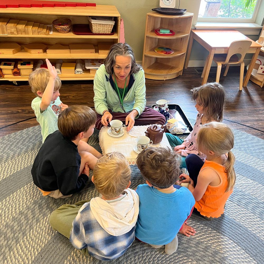 Children sit in a circle around a teacher at a table, sharing a small activity in a classroom.