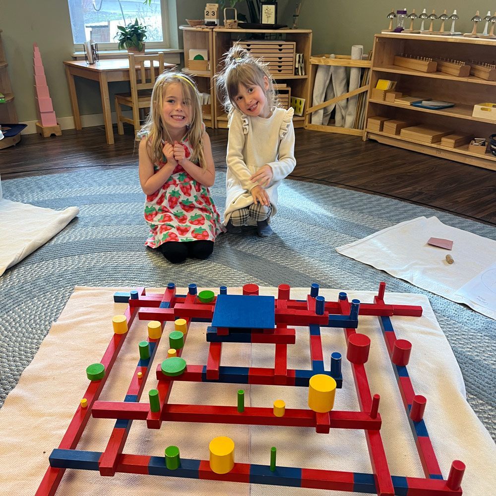 Two children kneel on a classroom rug behind a colorful block maze on the floor.