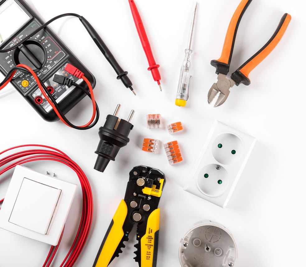 Herramientas y componentes eléctricos dispuestos sobre una superficie blanca, incluyendo un multímetro, alicates y cables.