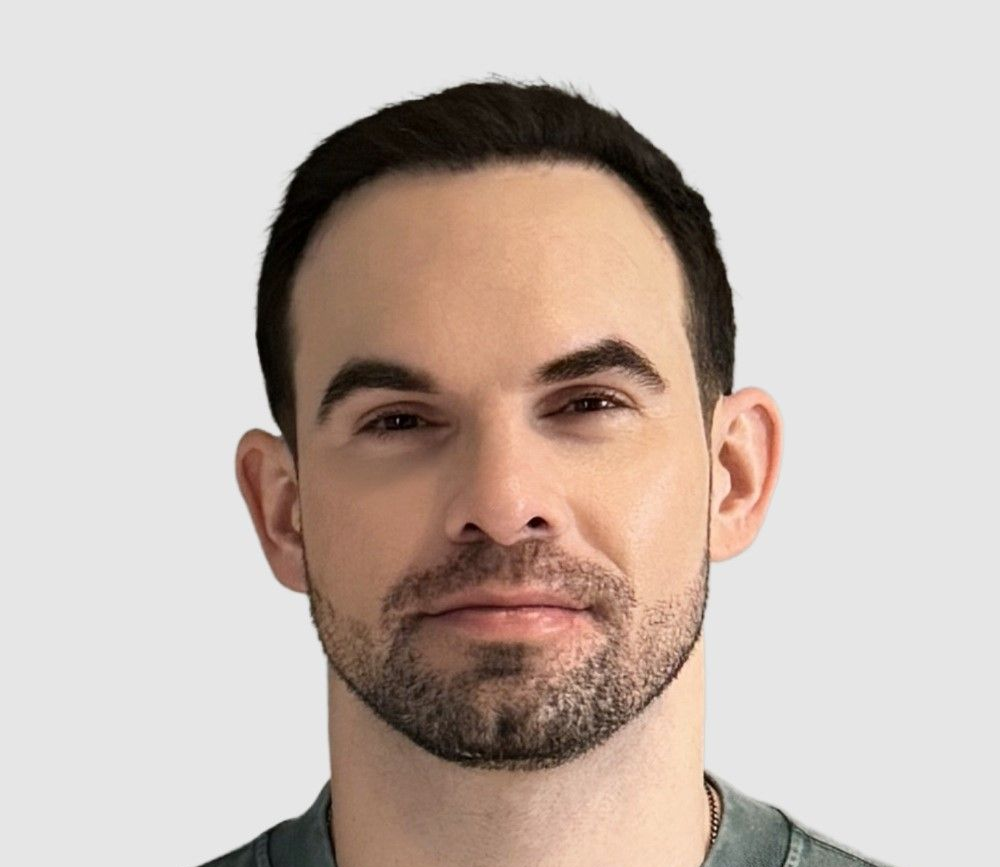 Man with dark hair and a beard against a light gray background, looking forward.