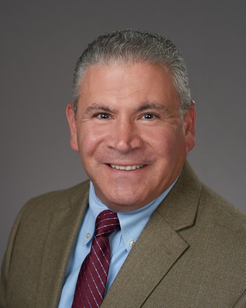 Man in a brown suit smiles, wearing a blue shirt and maroon tie, against a gray background.