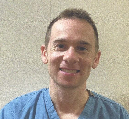 Man in blue scrubs smiling, in front of a neutral-toned wall.