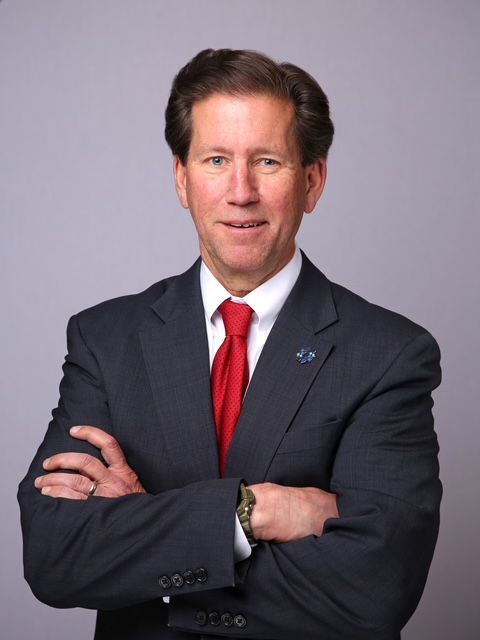 Man in a dark suit with a red tie and crossed arms, smiling at the camera.