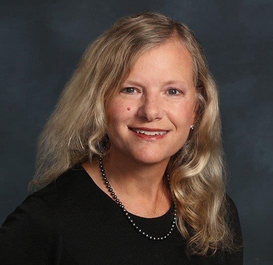 Woman with long blonde hair, smiling at the camera, wearing a black top and beaded necklace.