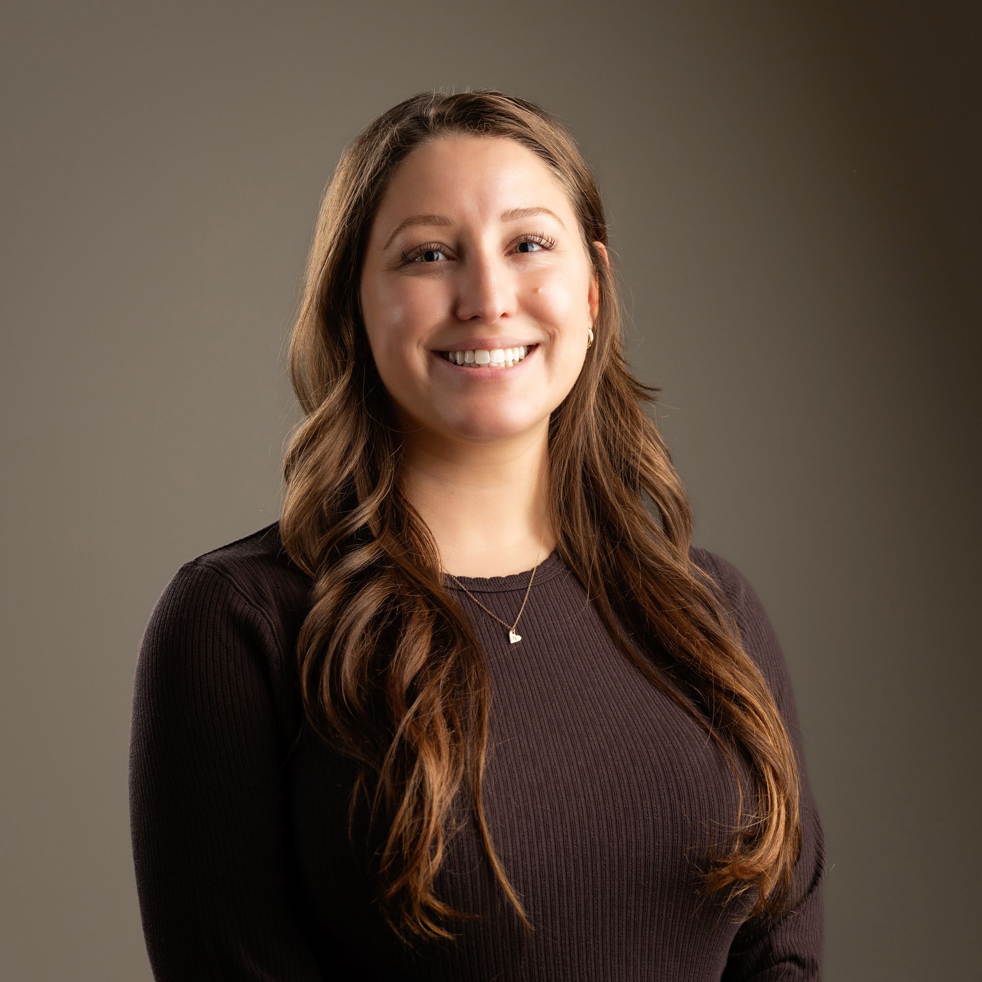 Woman smiling, wearing a dark sweater, with long brown hair, posing in front of a gray backdrop.