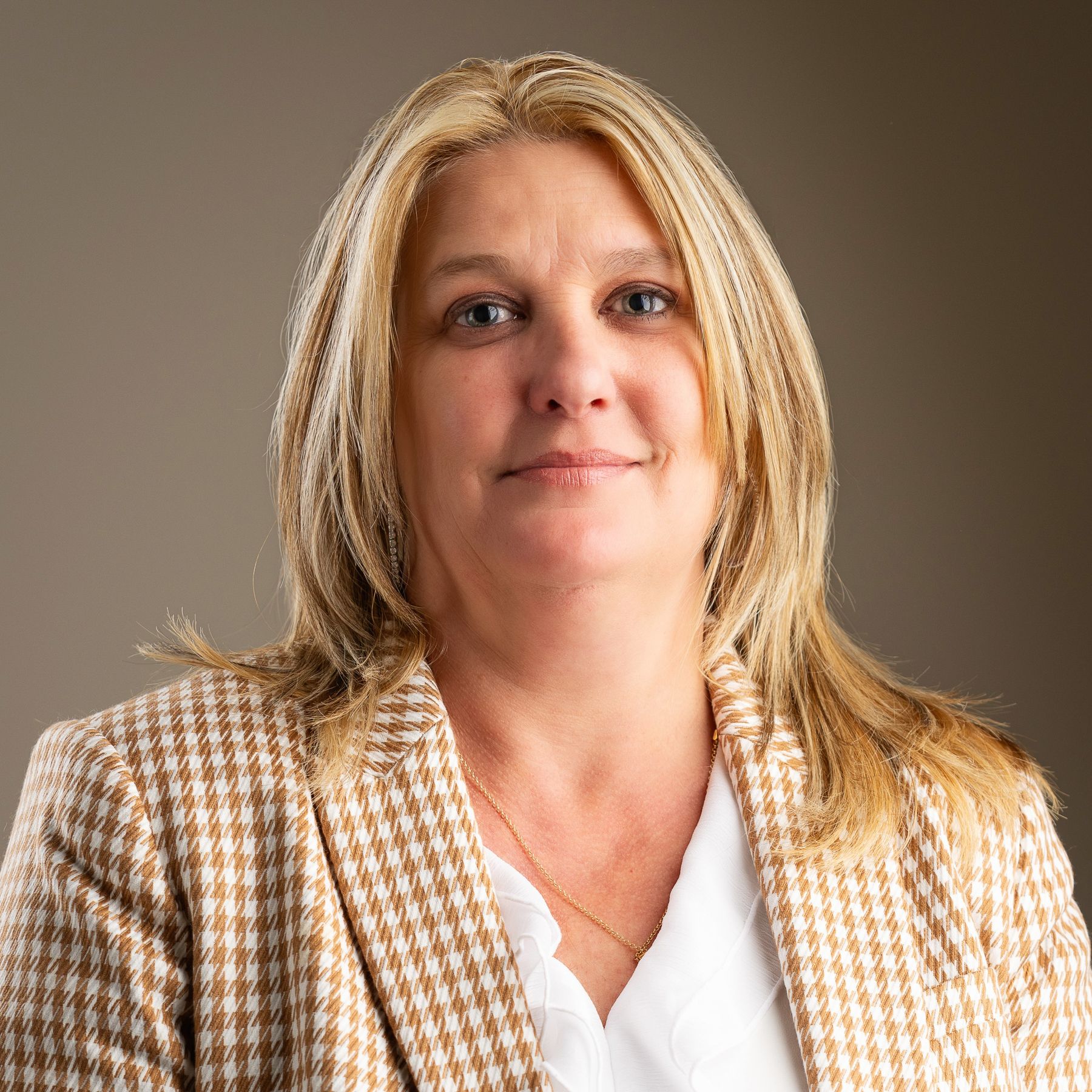 Woman with blonde hair wearing a white shirt and brown patterned blazer, looking at the camera.