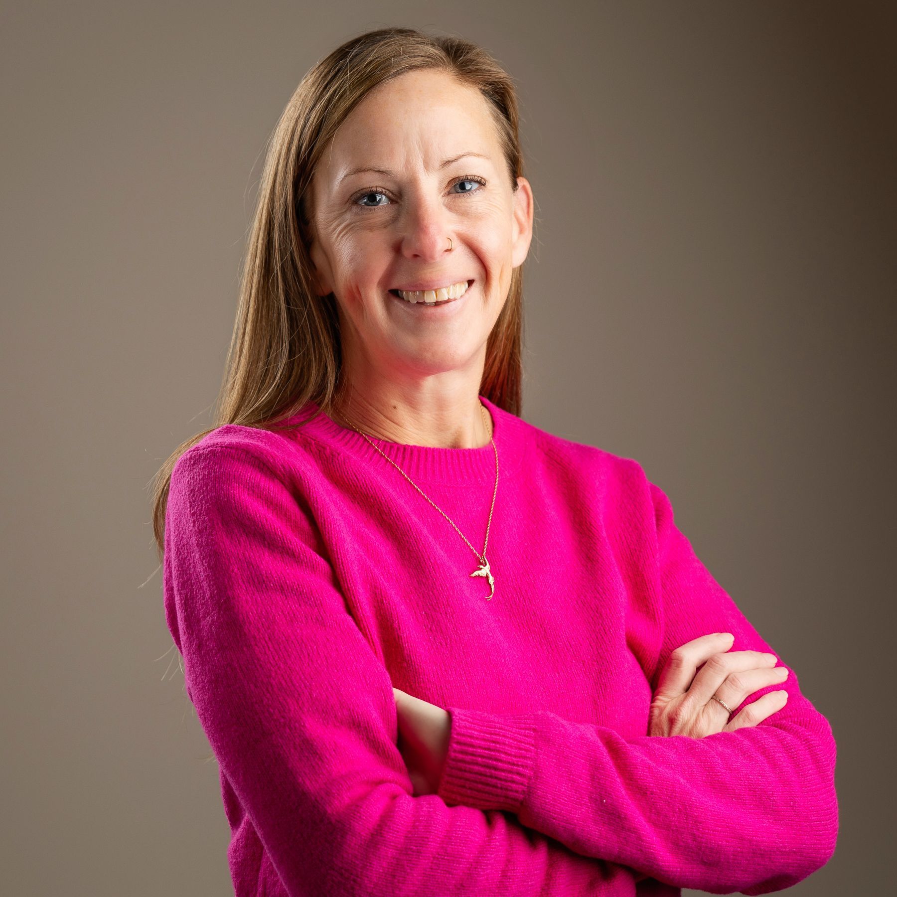 Woman wearing a pink sweater smiles, arms crossed, set against a neutral backdrop.