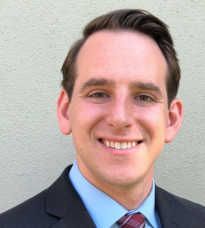 Smiling man in a suit and tie, blue shirt, against a light green wall.