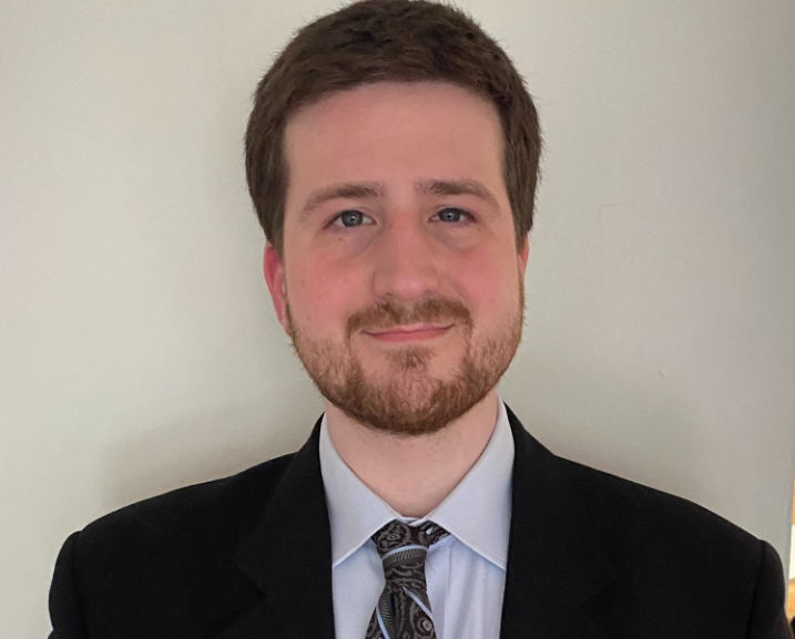 Man in black suit and tie smiles at the camera against a plain, light-colored wall.