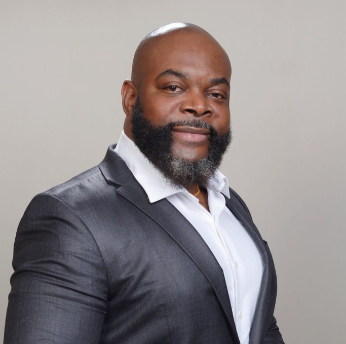 Man in a suit smiles, dark skin, bald head, full beard, white shirt, studio backdrop.