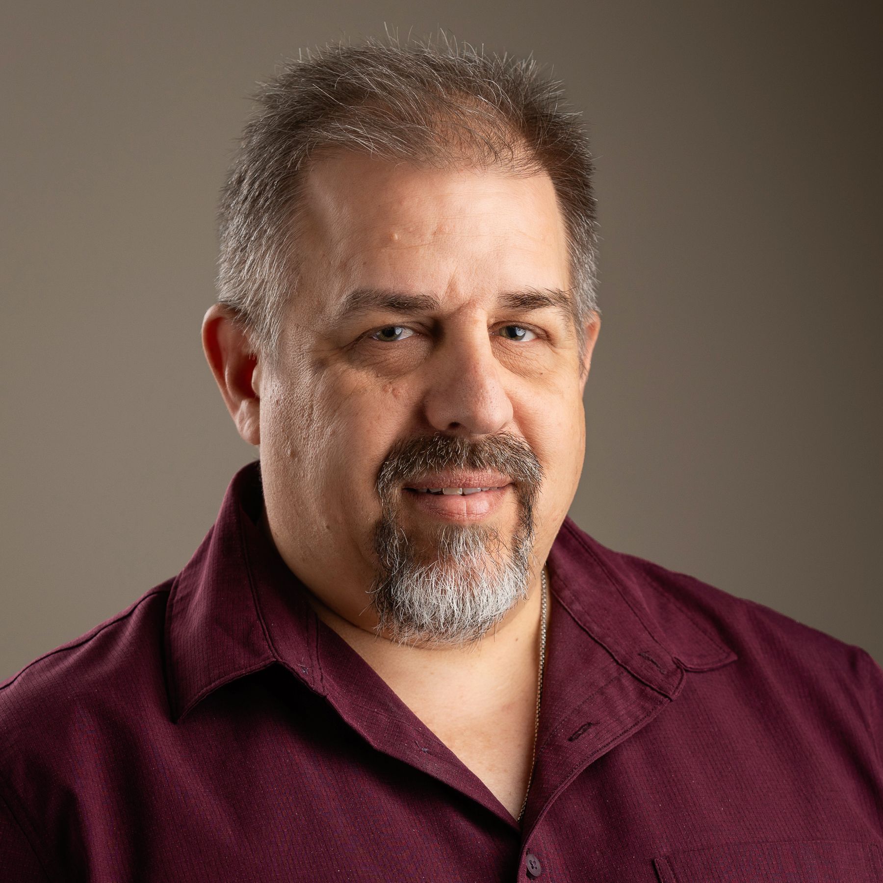 Man with short graying hair and beard smiles, wearing a burgundy button-up shirt against a neutral background.