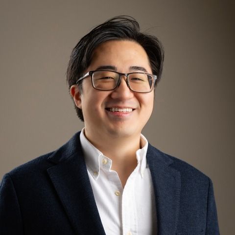 Man in glasses, smiling, wearing a navy blazer and white shirt, against a neutral background.