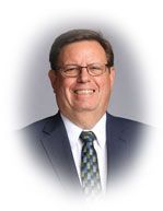 Man in glasses, suit and tie, smiling, against a blurred background.
