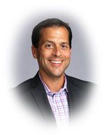 Man smiling, wearing a blazer over a plaid button-down shirt; a gray gradient backdrop.