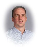 Man wearing a light blue striped button-down shirt smiles, soft background.