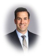 Man in suit smiling, with a light blue shirt and grey tie against a gradient background.