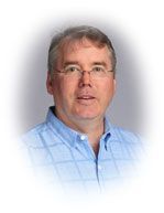 Man in glasses and blue shirt smiles, headshot with blurred background.
