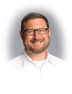 Man with glasses and beard smiles, wearing a white shirt; against a gray background.