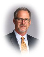 Man in a suit and tie, wearing glasses, smiling slightly, in a studio setting with a white vignette.