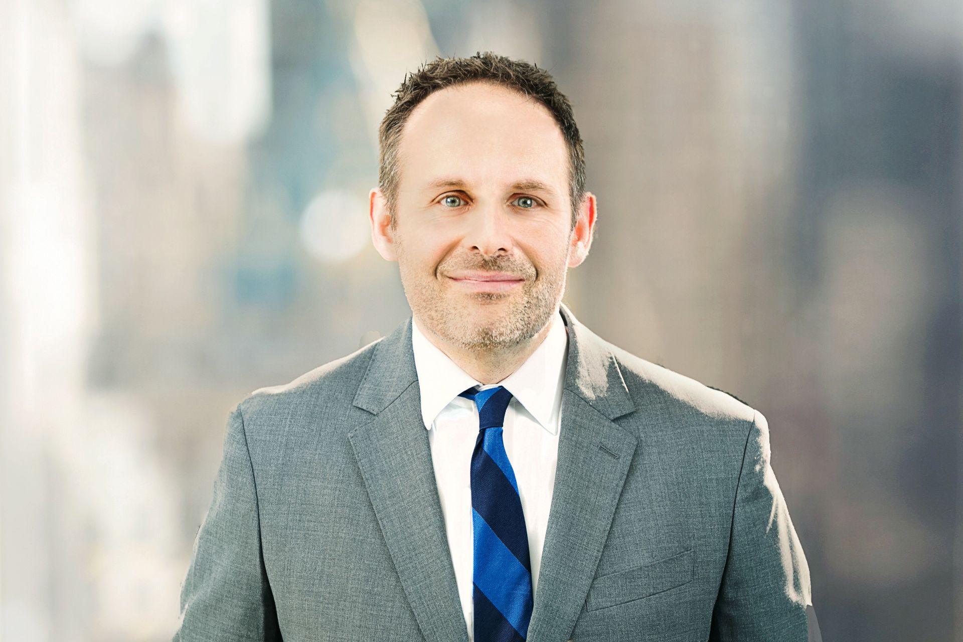 Man in a gray suit with a blue tie, smiling, indoors with a blurred city backdrop.
