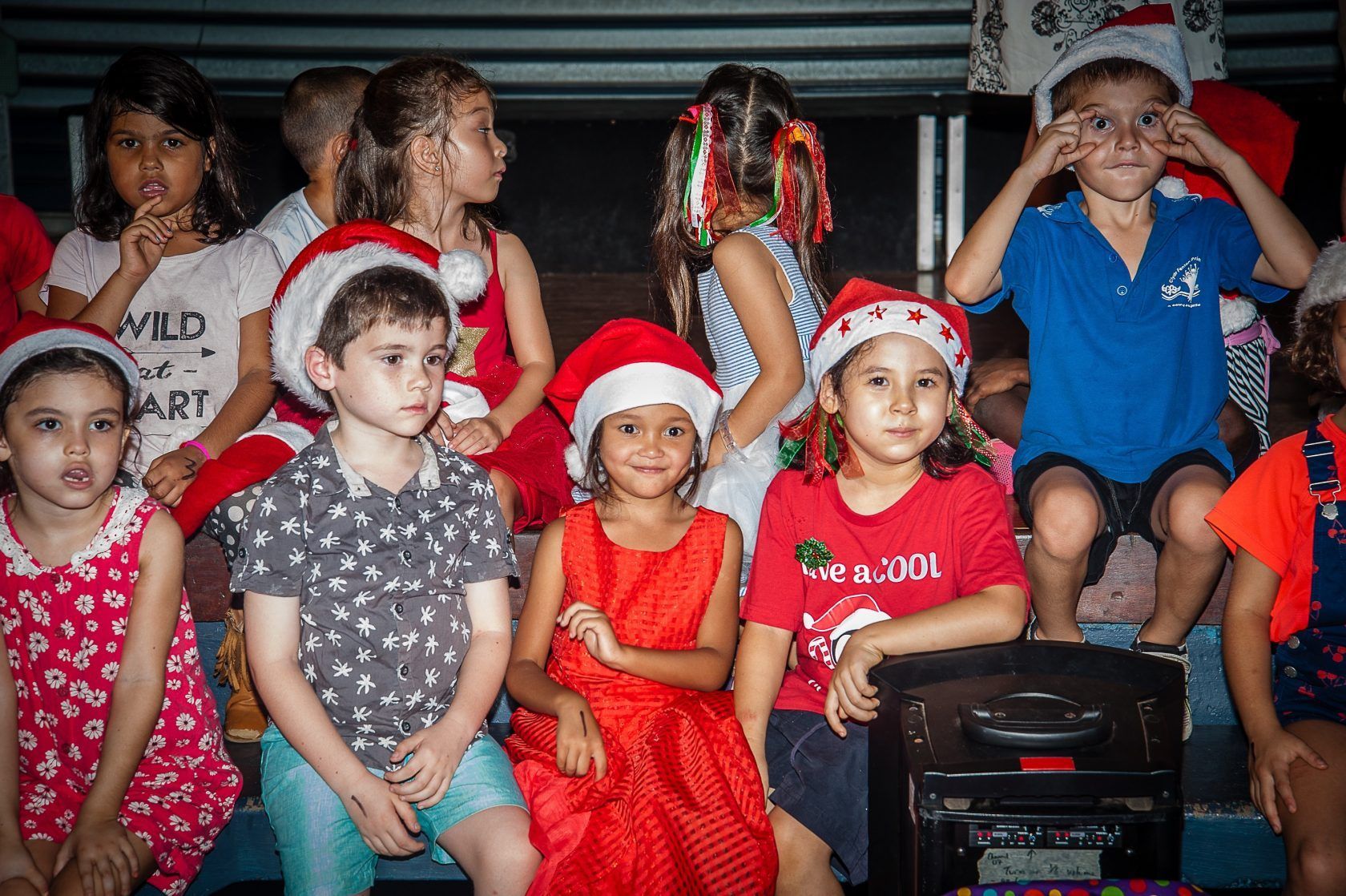 A group of children wearing santa hats are sitting on a stage.
