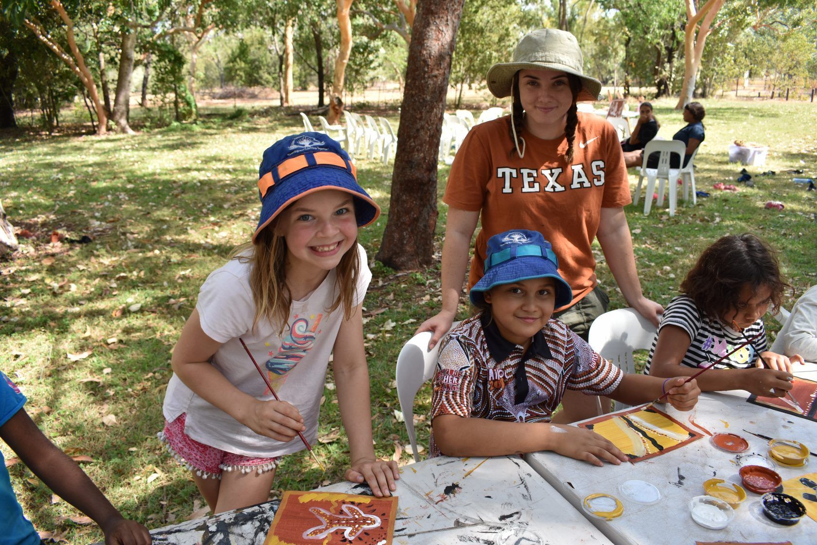 A group of children are sitting at a table with a woman wearing a texas shirt.