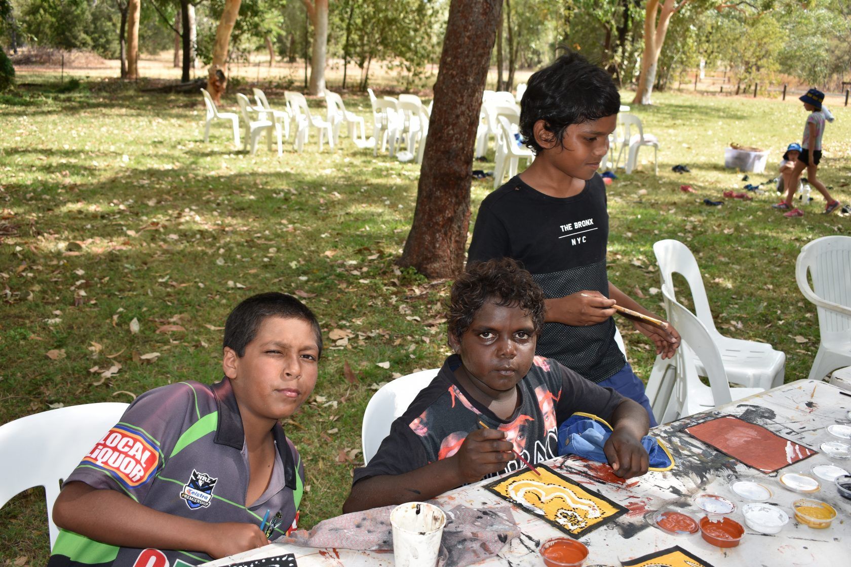 Three young boys are sitting at a table in a park painting.