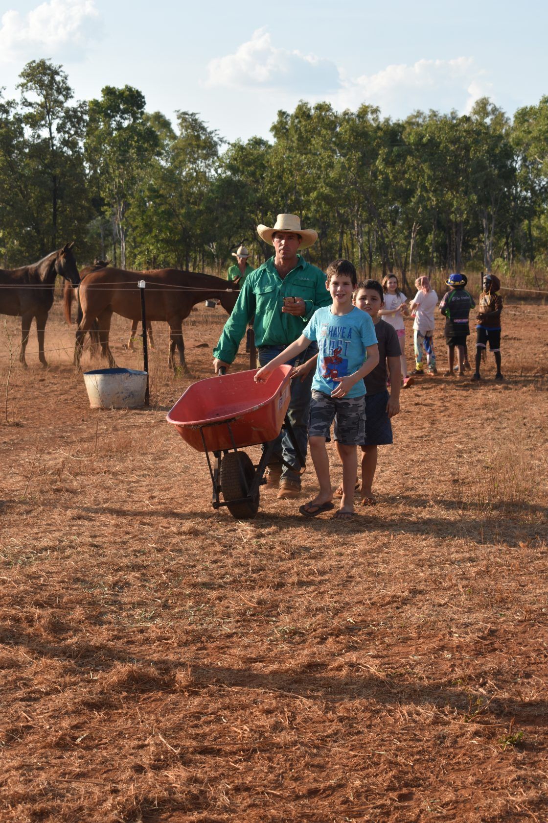 A man pushing a wheelbarrow in a field with horses in the background