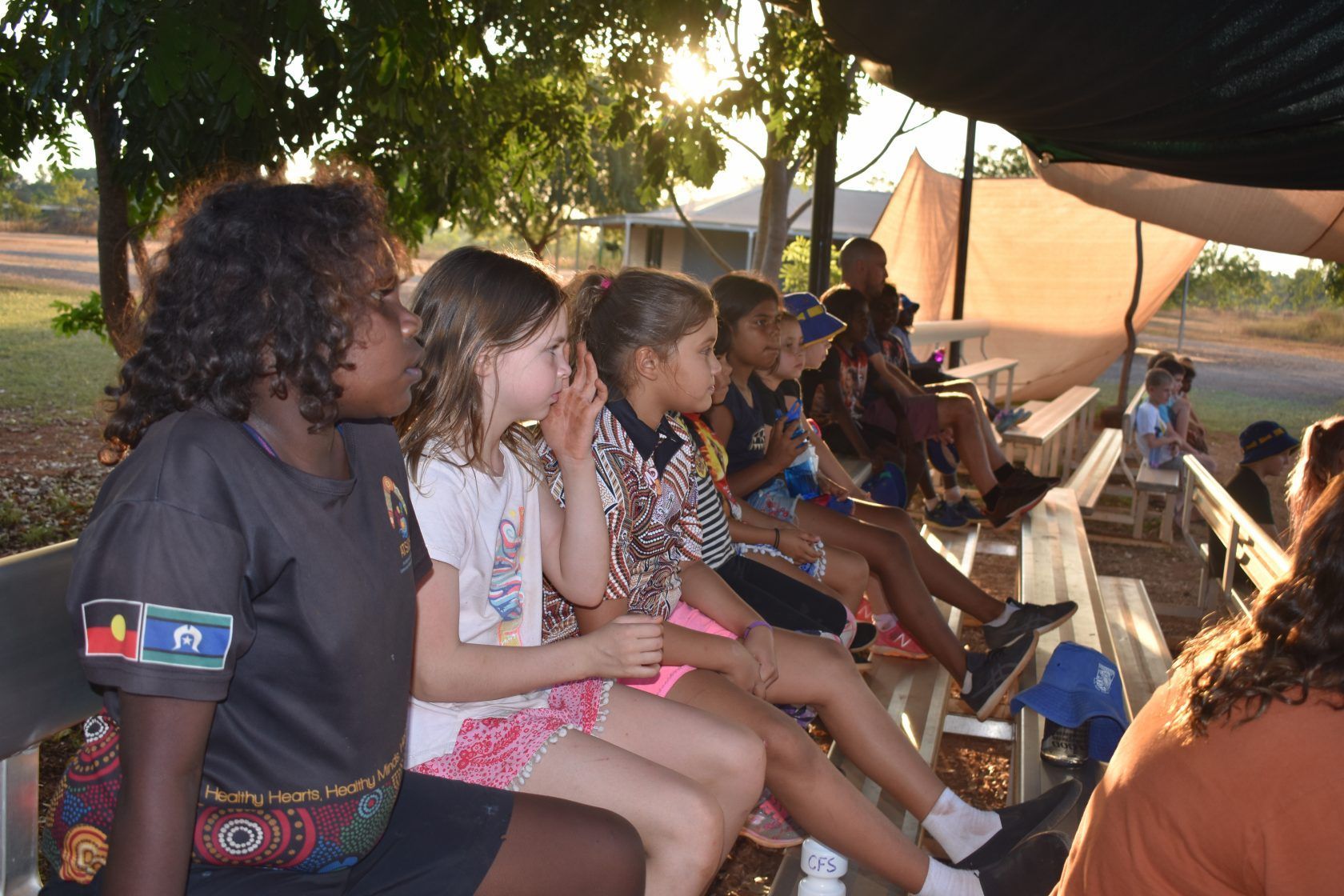 A group of children are sitting on a bench under an umbrella.