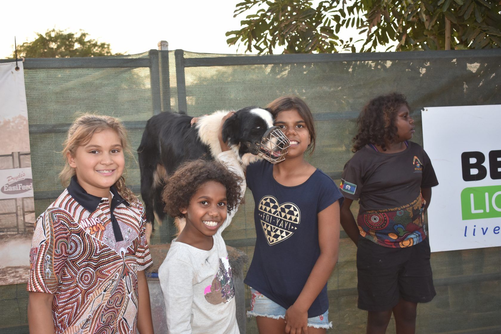 A group of young girls standing next to a dog in front of a sign that says be live