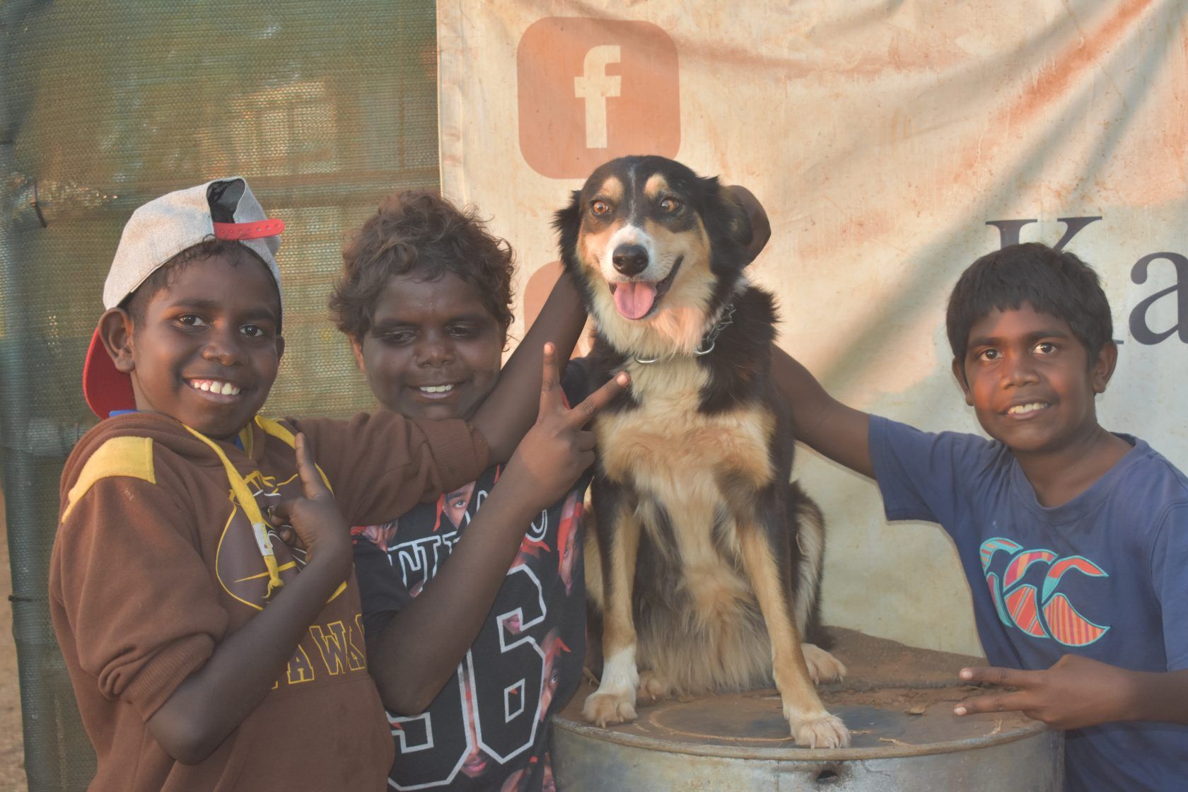 Three young boys are posing with a dog in front of a facebook banner