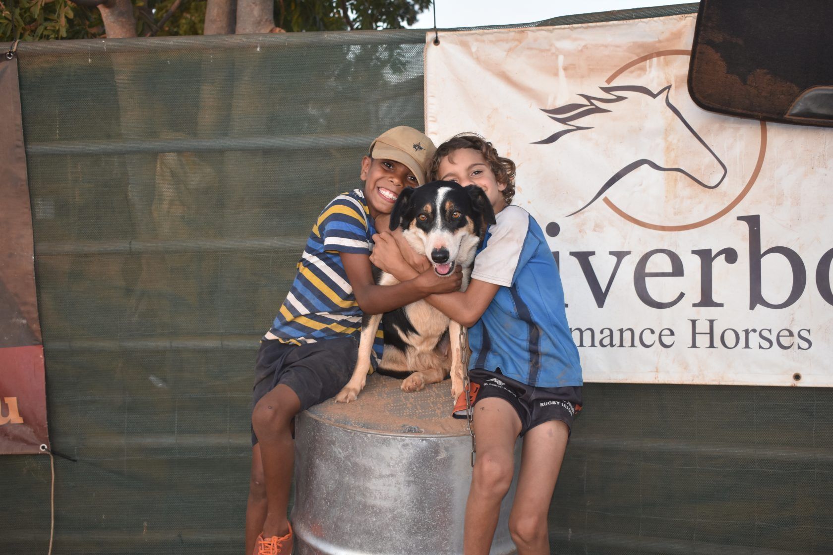 Two boys hugging a dog in front of a sign that says verbo