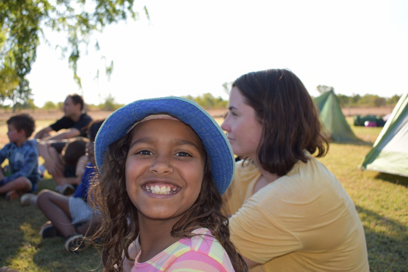 A young girl wearing a blue hat is smiling for the camera while sitting in the grass.