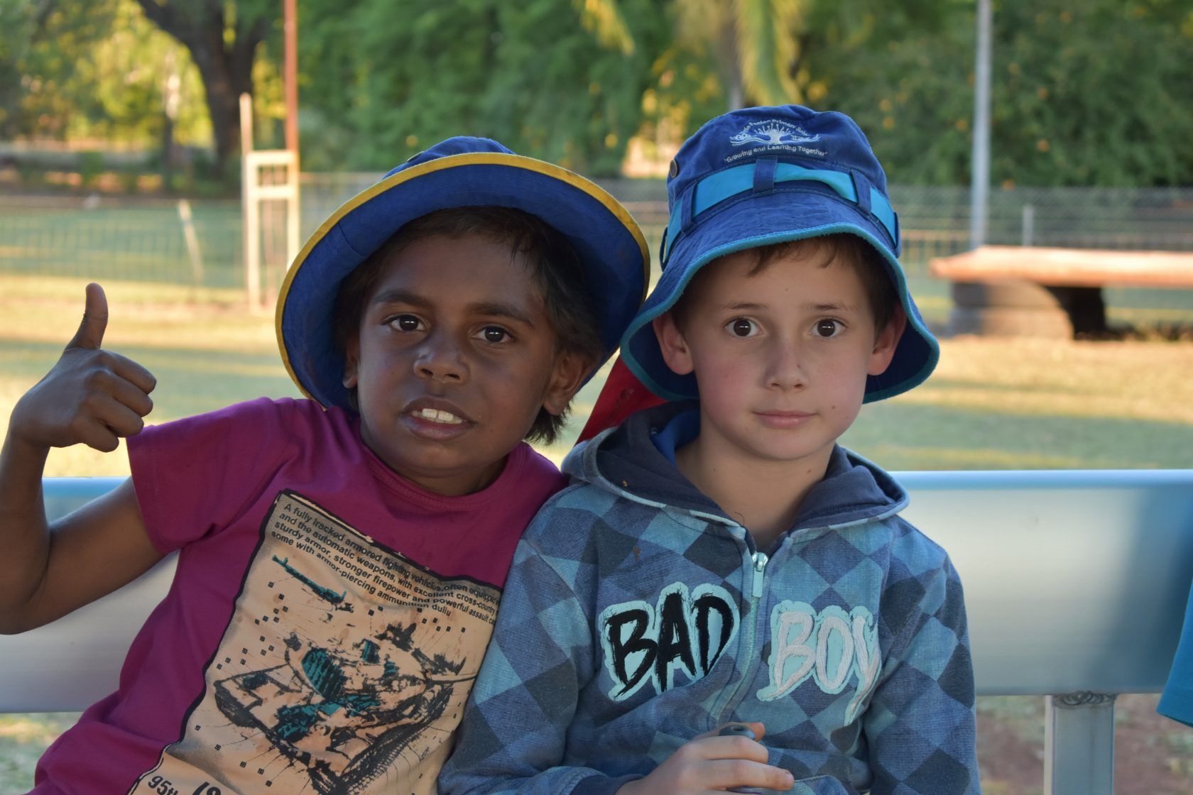 Two young boys wearing hats are sitting on a bench giving a thumbs up.