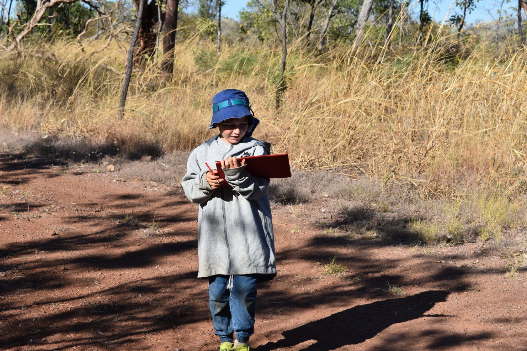 A young boy is standing on a dirt road holding a clipboard.