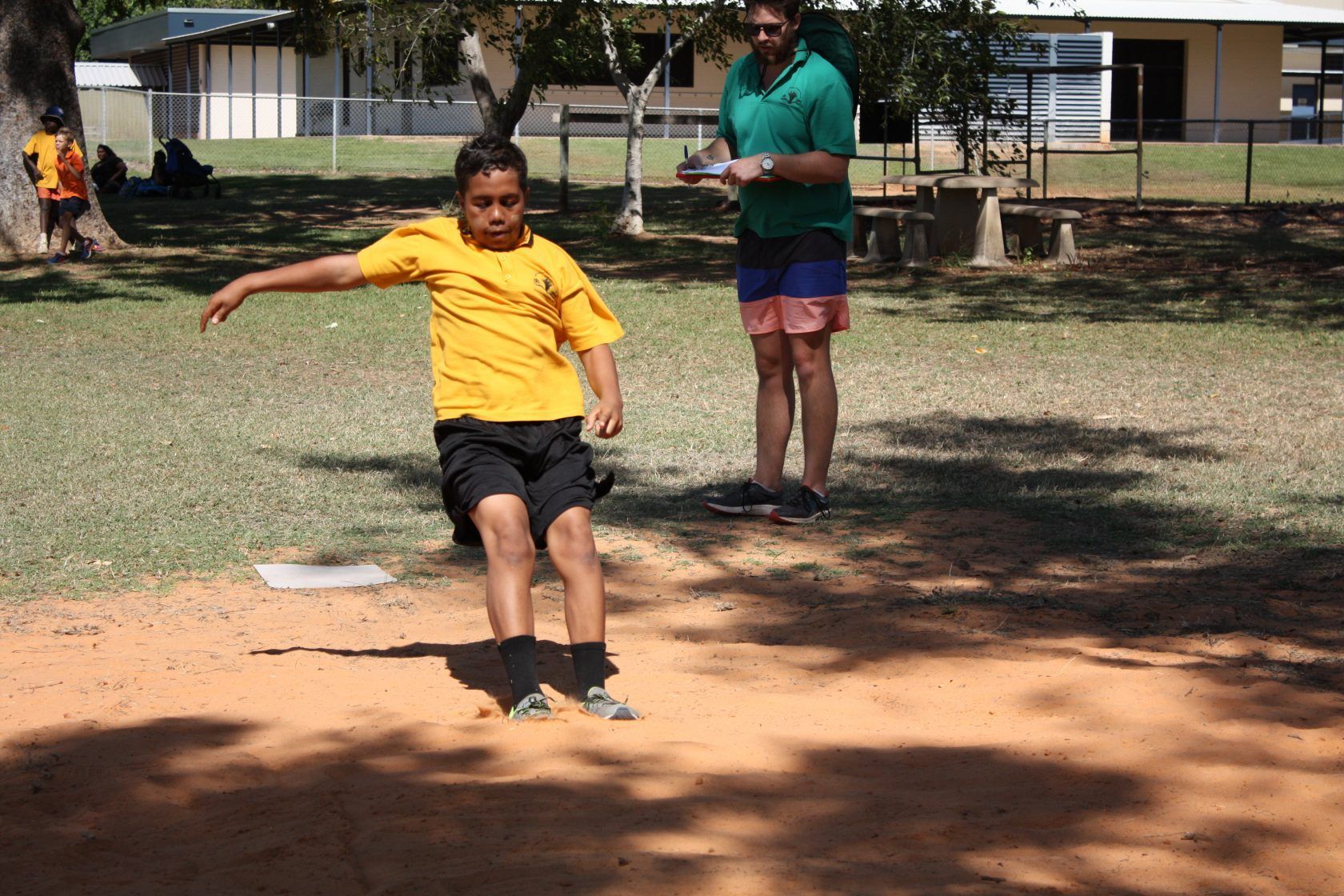 A young boy is jumping in the dirt while a man watches.