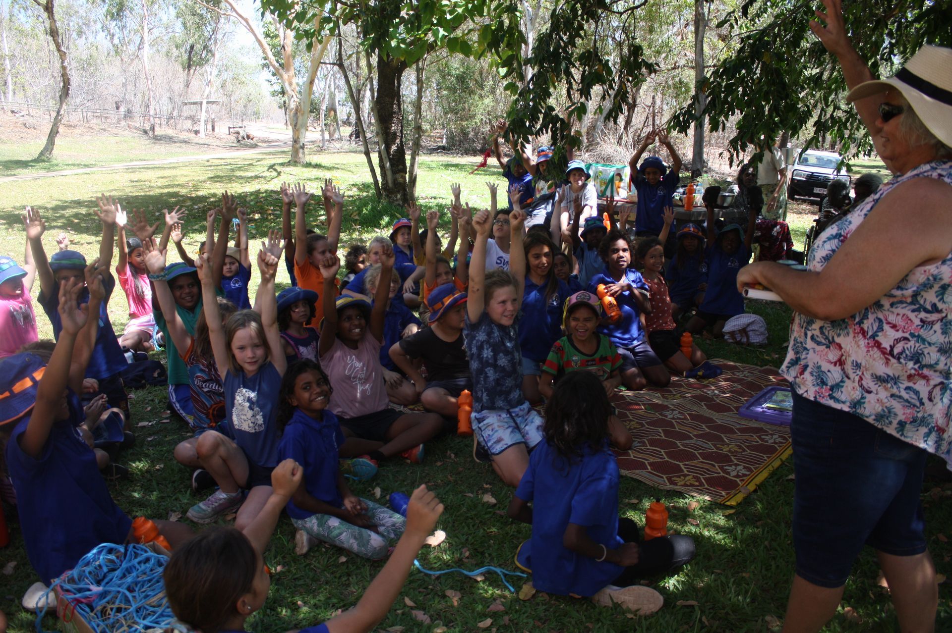 A group of children are sitting on the grass with their hands in the air.