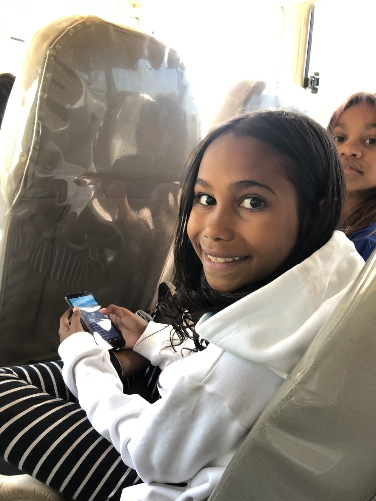 A girl is sitting in the back seat of a bus holding a cell phone.