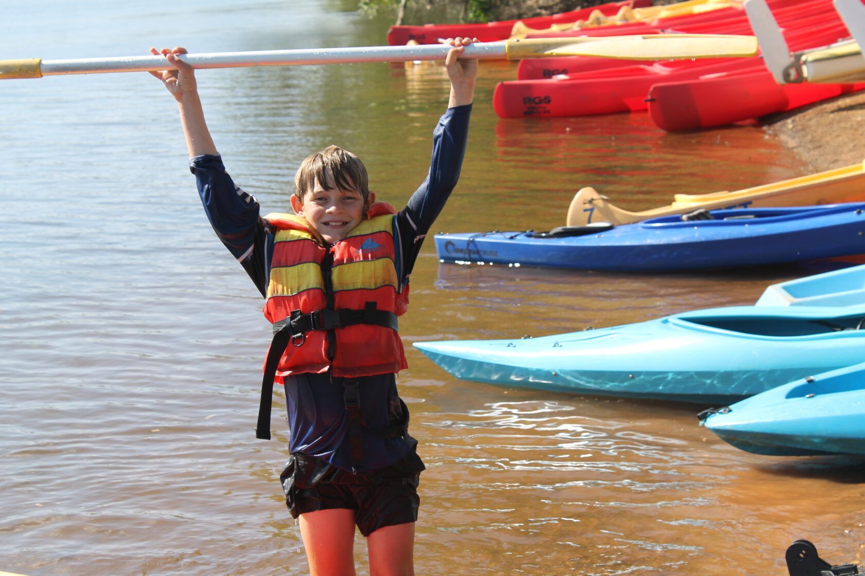 A boy in a life jacket is holding a paddle over his head in the water