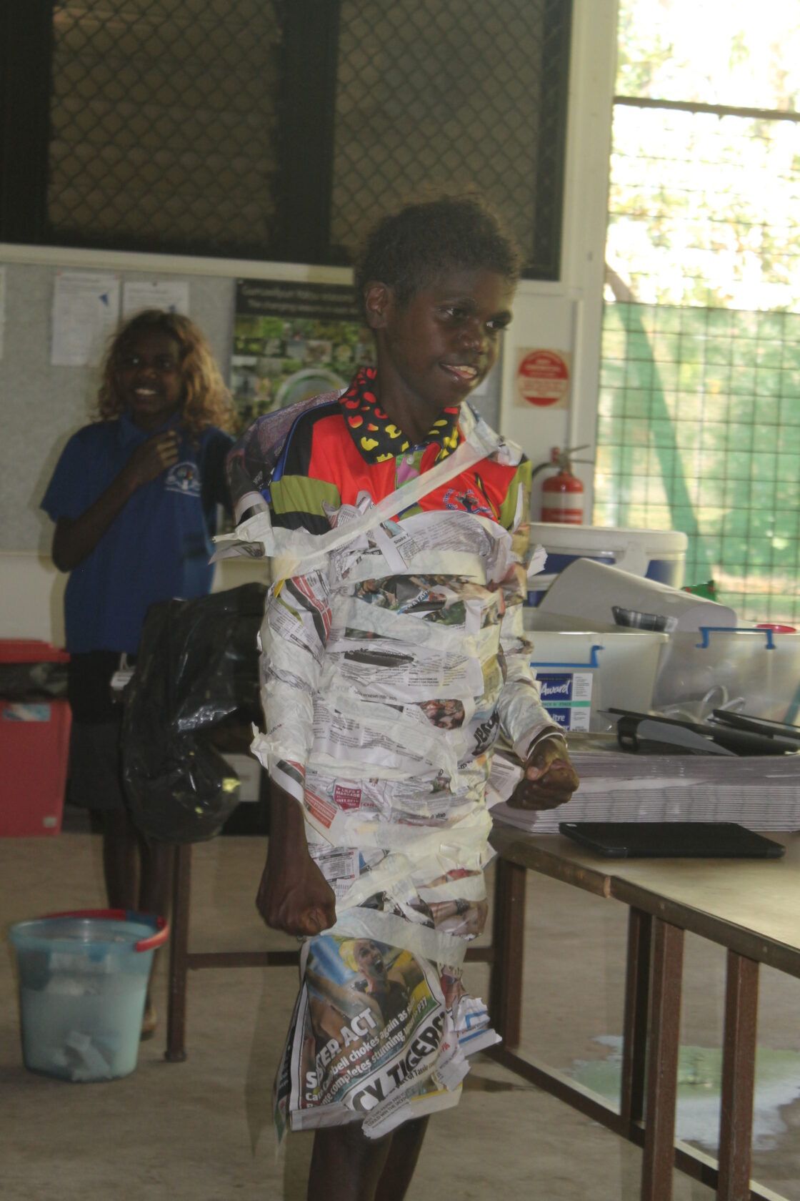 A young boy is wrapped in newspaper in a classroom.