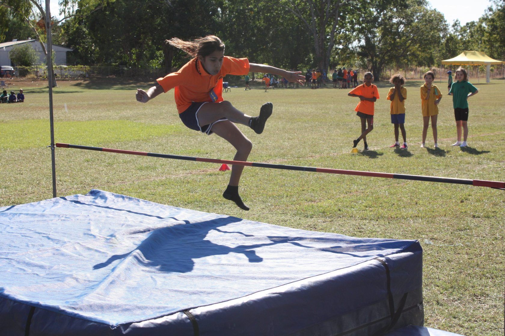 A girl in an orange shirt is jumping over a high jump bar
