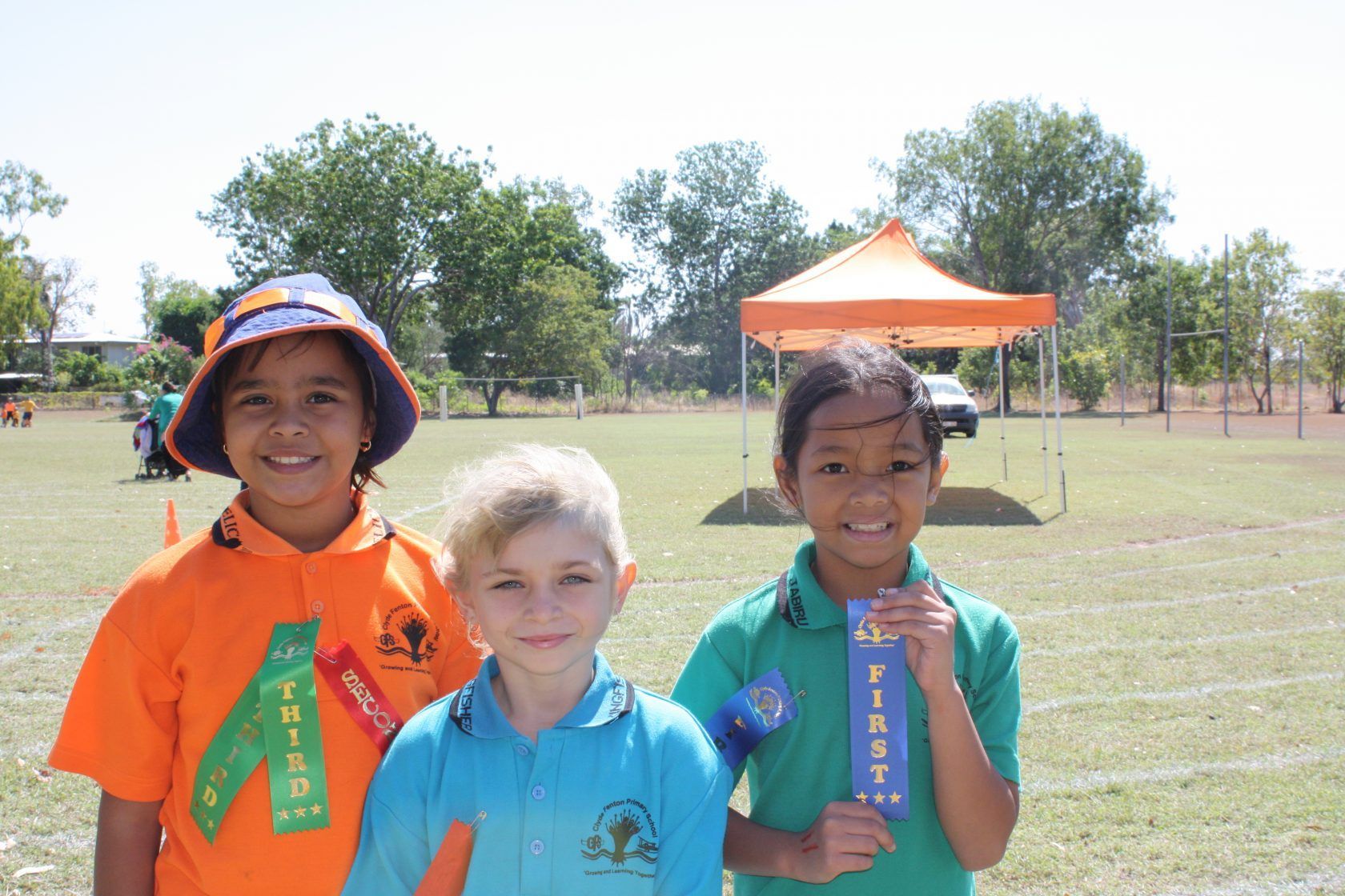 Three children are posing for a picture and one of them is holding a ribbon that says first