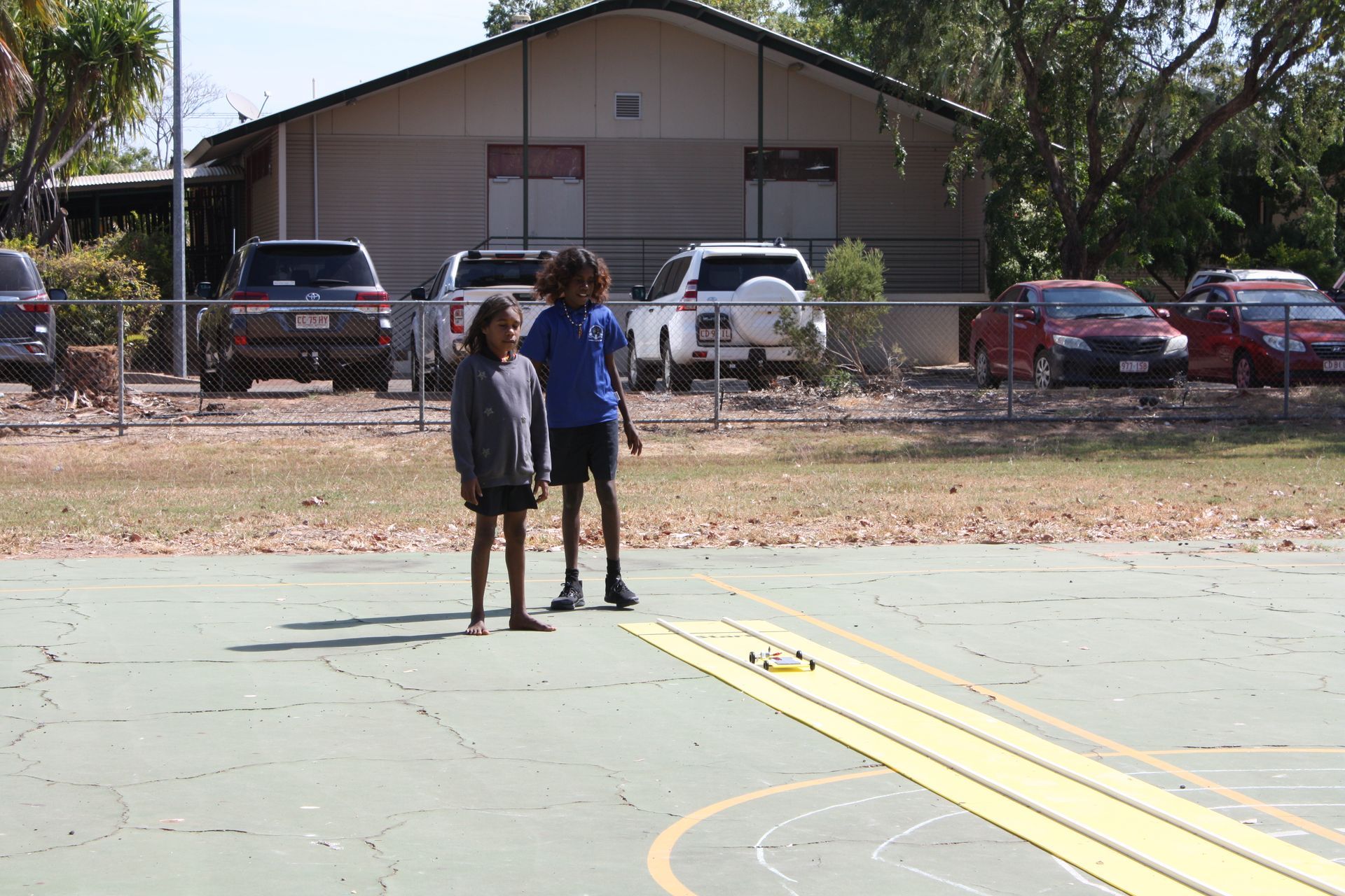 Two children are standing on a basketball court in front of a house.