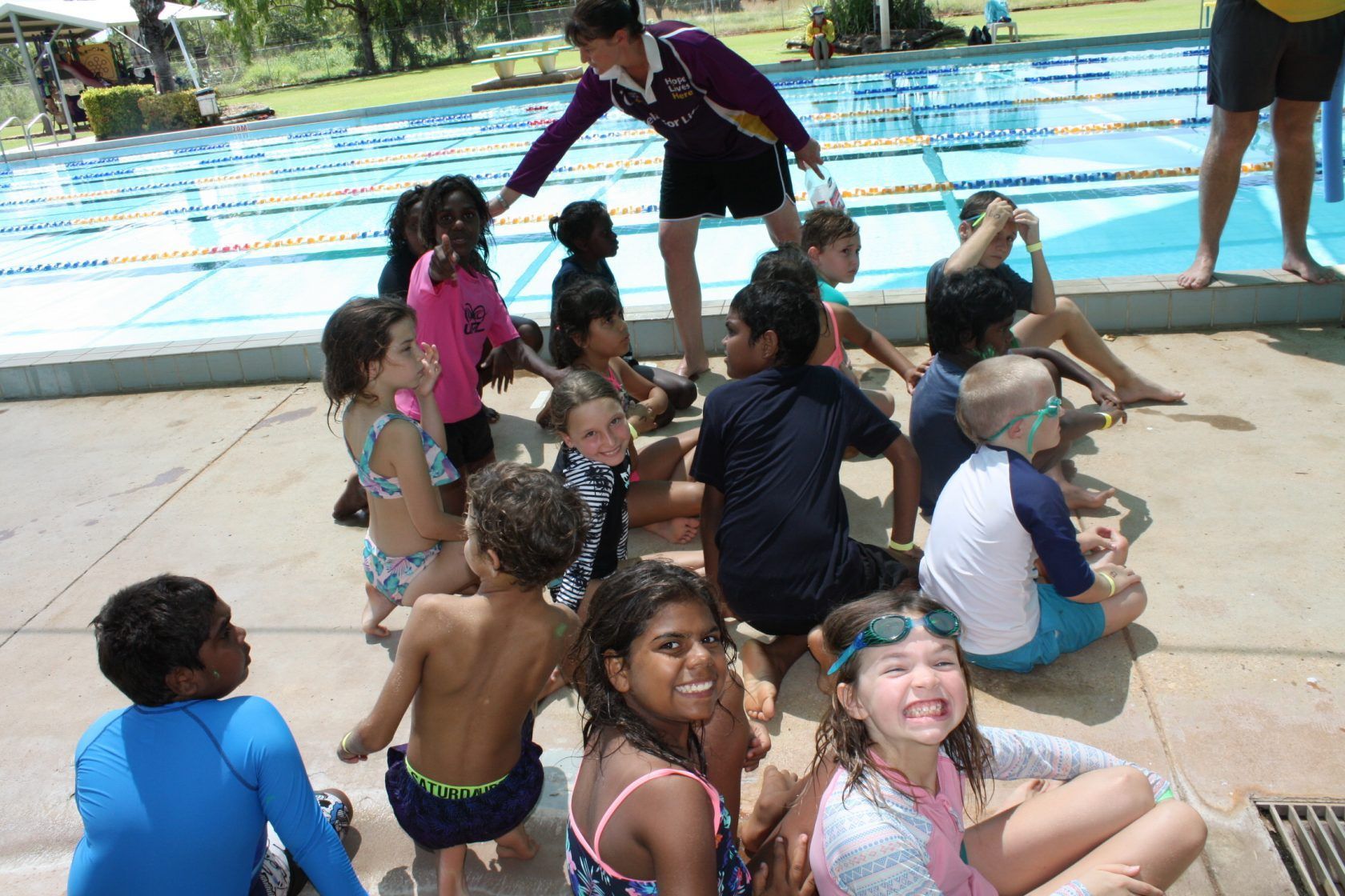 A group of children are sitting on the ground near a swimming pool