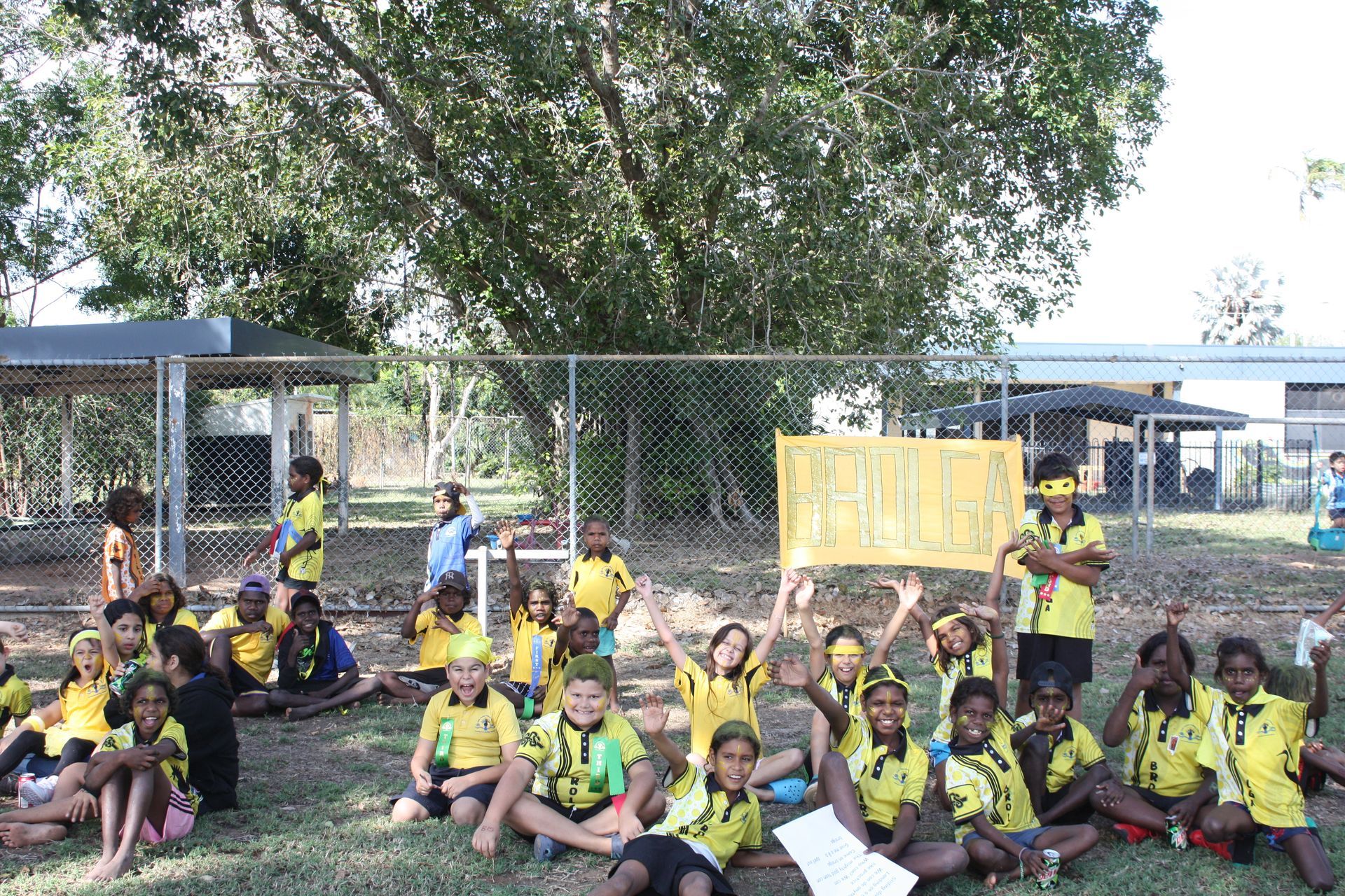 A group of children are sitting on the grass in front of a fence.