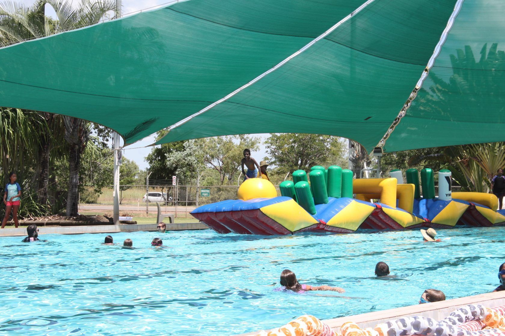 A group of people are swimming in a pool under a green umbrella.