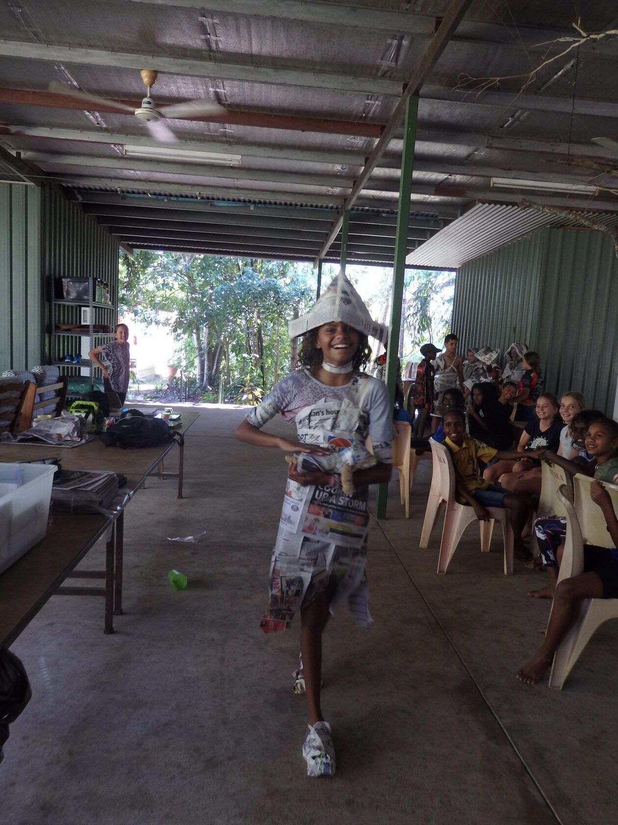 A young girl is standing in a room wearing a hat made out of newspapers.