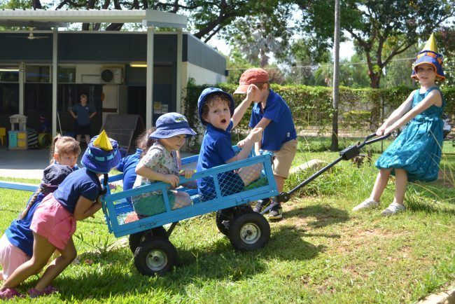A group of children are pushing a blue wagon in the grass.