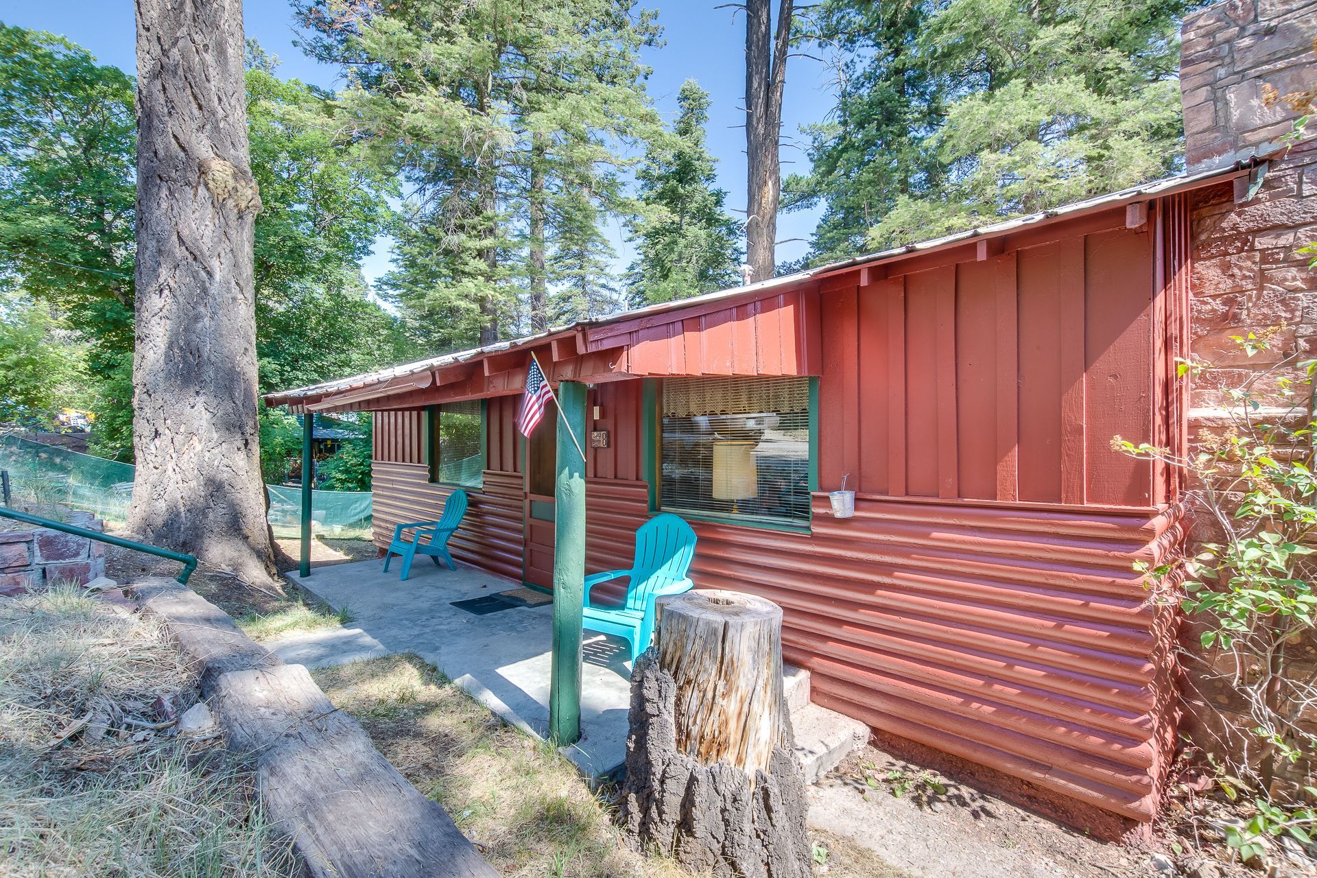 A red log cabin with blue chairs on the porch surrounded by trees.