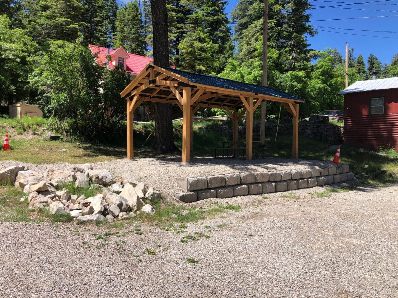 A wooden pavilion is sitting on top of a gravel driveway next to a stone wall.